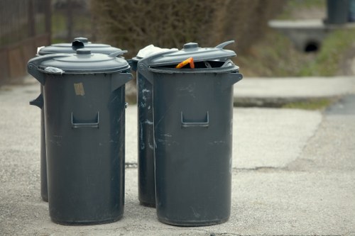 Two dark grey plastic wheelie bins with closed lids are positioned side by side on a paved street or driveway area, with one bin's lid slightly ajar revealing an orange waste item inside. The bins are situated outdoors, with a blurred background showing a grassy or natural environment and distant trees. The surface around the bins appears to be concrete or asphalt, typical of residential or urban areas in Golders Green. These bins are part of waste management and rubbish removal services provided by Office Clearance Golders Green, emphasizing their role in local rubbish collection efforts within the area. The lighting suggests an overcast day, with diffused natural light illuminating the scene evenly. The bins are clean, with a smooth finish, and are commonly used for household or commercial waste disposal, aligning with professional rubbish removal operations in the region.