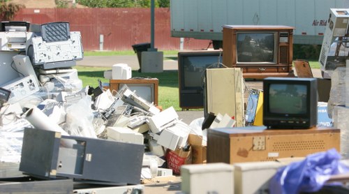 A large pile of discarded electronic waste and outdated electrical appliances, including old computer monitors, televisions, circuit boards, and various electronic components, is seen outdoors on a paved surface. The heap includes a mix of beige, black, gray, and brown devices, some stacked and leaning against each other, with plastic bags and packaging materials interspersed among them. In the background, a low red brick wall and a green metal fence are visible, with a grassy area and trees beyond, indicating an outdoor setting in or near Golders Green. The scene is illuminated by daylight, highlighting the textures of the aged plastic casings and metal finishes. This image, associated with rubbish removal services by Office Clearance Golders Green, illustrates the type of electronic waste typically handled during customer clearance projects in the local area, emphasizing the importance of proper disposal of outdated electrical items.