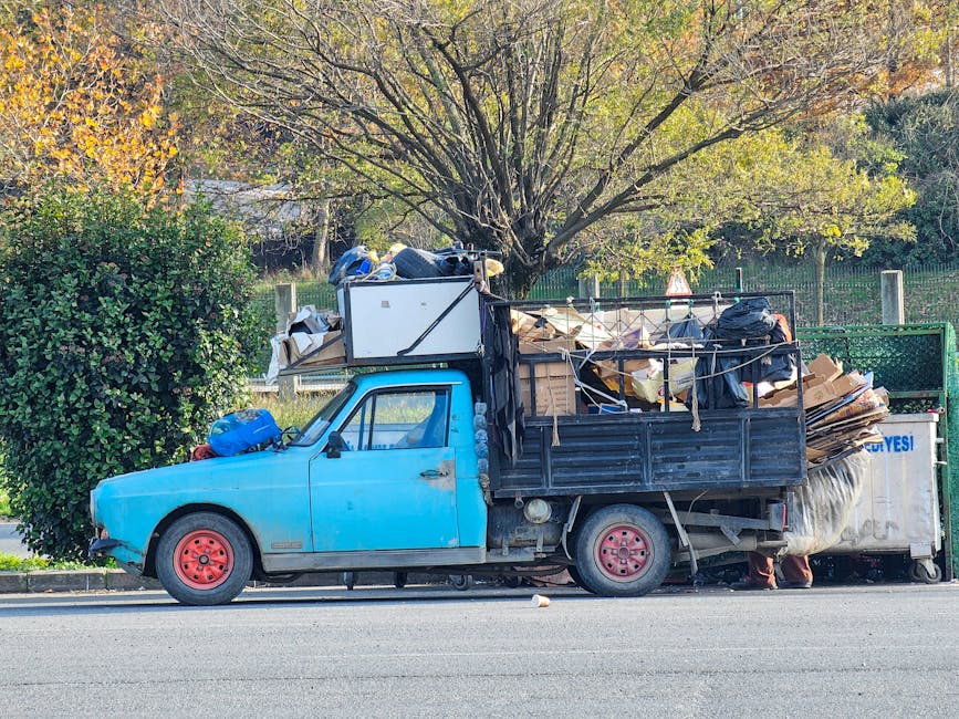 A small, older model blue flatbed truck parked alongside a paved street with only the driver’s side visible. The truck's exterior shows signs of wear, with faded and chipped paint, particularly on t